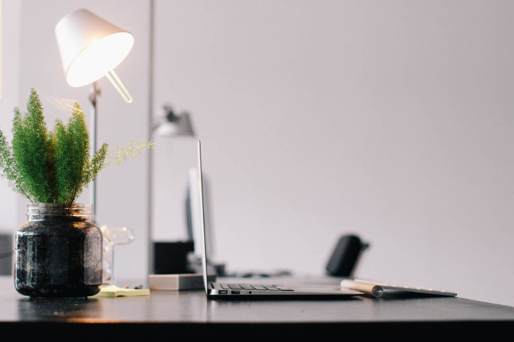 Zu sehen ist ein ordentlich aufgeräumter Schreibtisch, mit einem Laptop, eine Pflanze in einem Glas Topf und einer Hell Leuchtenden Schreibtischlampe.